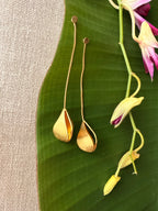 A pair of gold tones dangle earrings with petal pendants, displayed on a large green banana leaf next to pink and yellow orchid flowers on a beige linen surface.