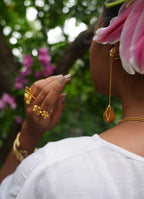 A woman shown from the back wearing a long gold dangle earring, a gold necklace, and gold rings, with a pink flower tucked into her hair against a blurred green outdoor background.