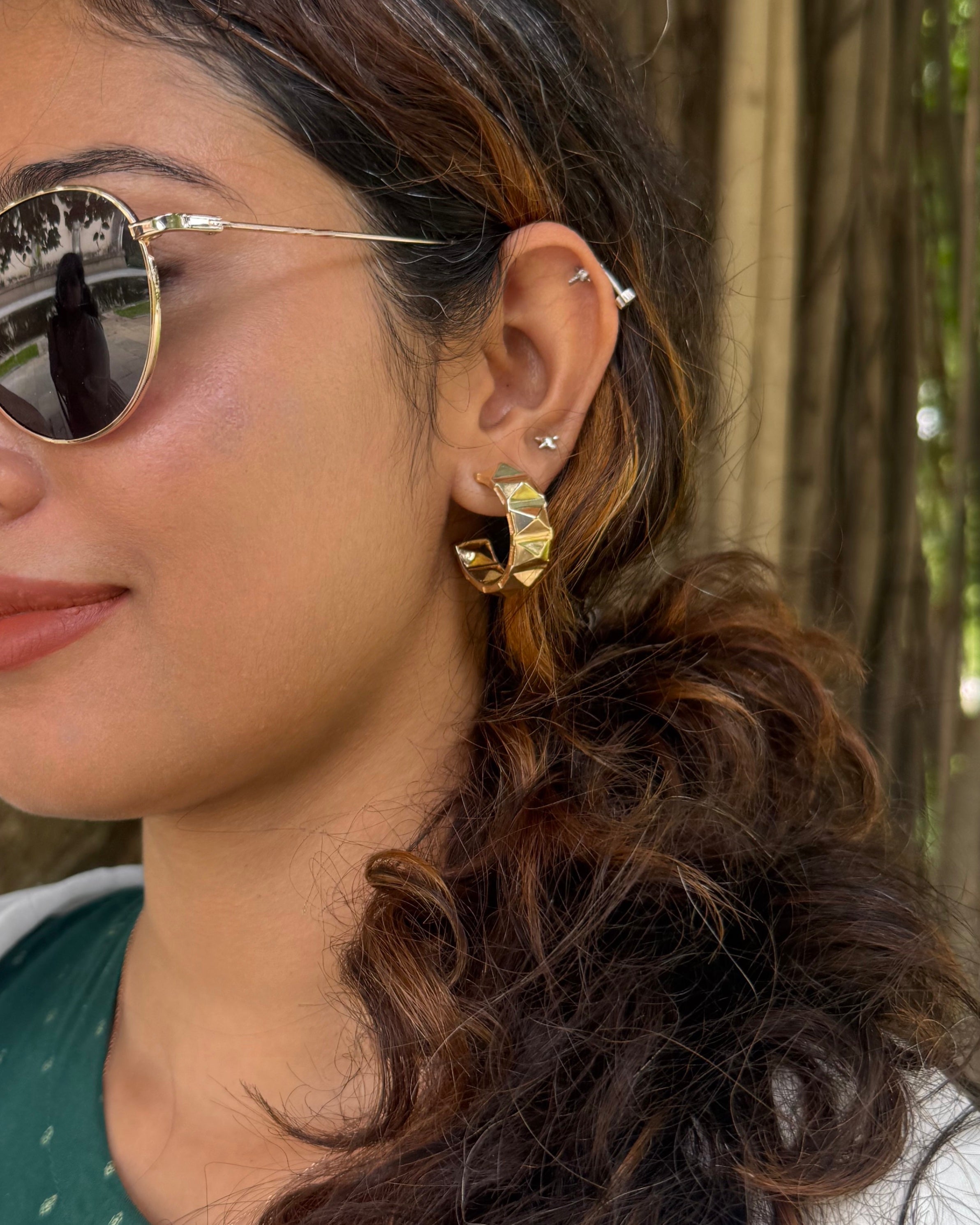Close-up of a woman wearing gold plated geometric hoop earrings with a blurred natural background