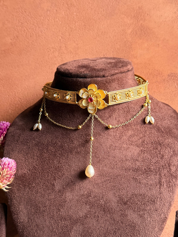 Nargis Choker displayed on velvet bust against terracotta wall, gold filigree band with central flower and ruby-coloured stone, pearl drop chains cascading down, dried pink flowers beside it.
