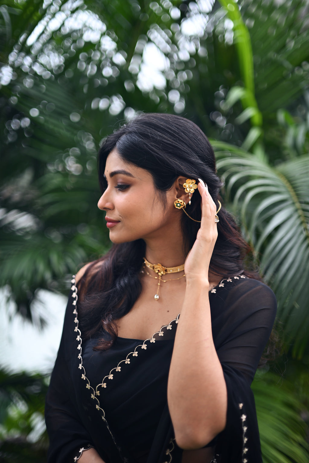 Side profile of woman wearing Nargis Ear Cuffs and Nargis Choker, black embroidered outfit, hand raised to ear, lush tropical greenery in background, soft natural light