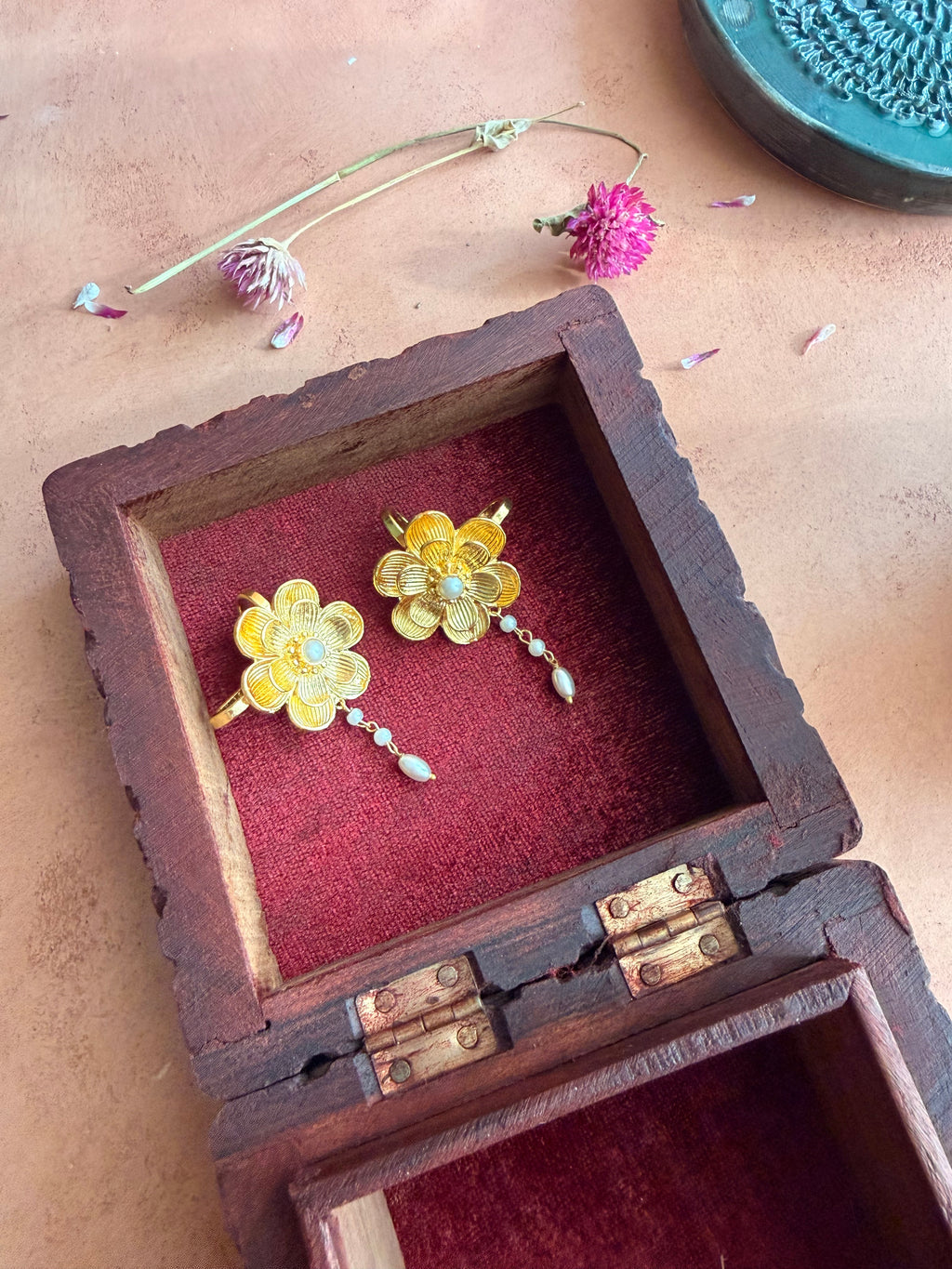 Nargis Ear Cuffs nestled inside an antique wooden box with red velvet lining, gold flower and pearl drop visible, dried flowers and petals on terracotta surface around it