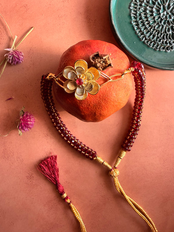 Nargis Mala garnet beaded necklace with gold flower pendant draped over a pomegranate, styled with dried flowers and a green plate.