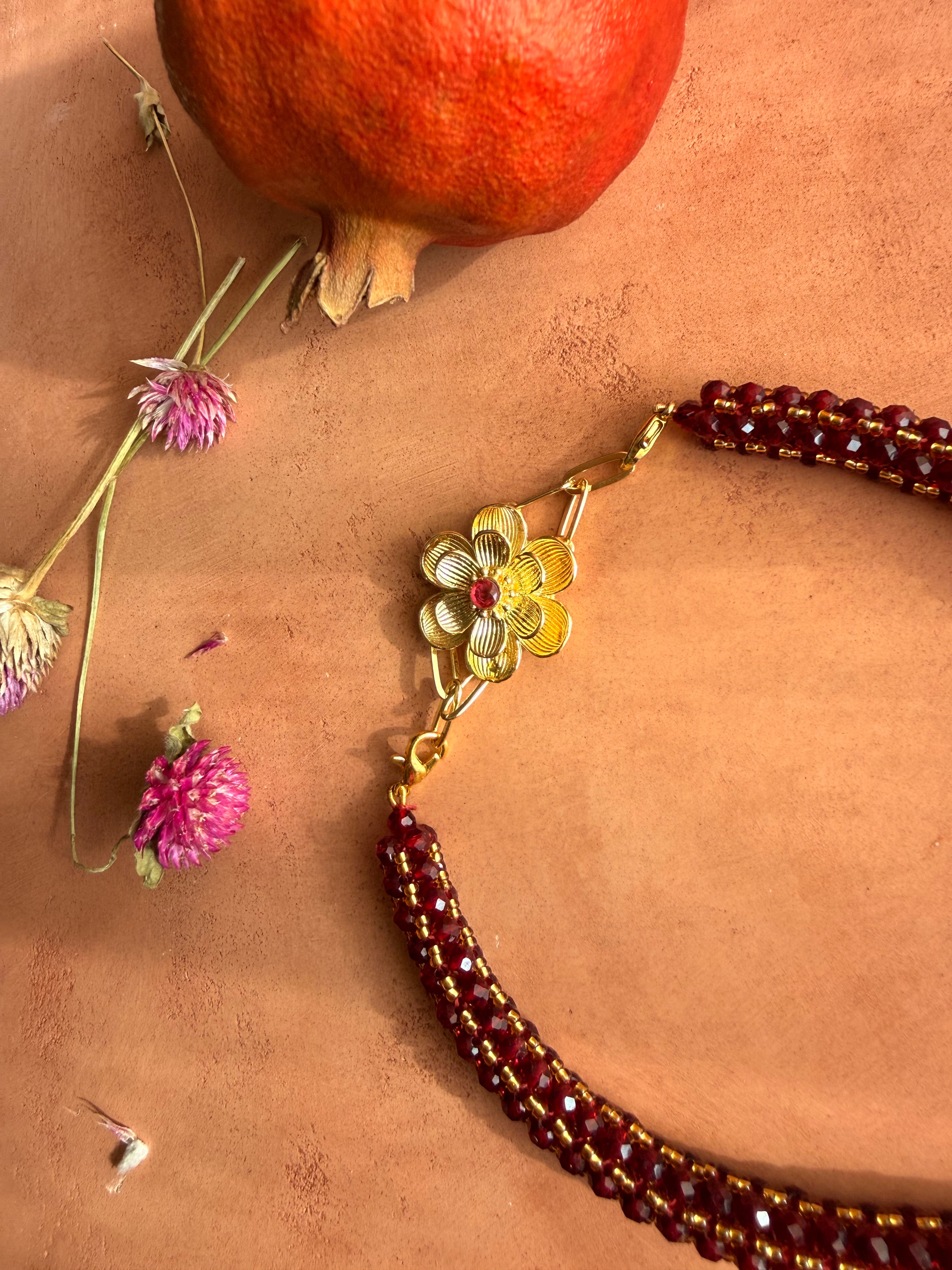 Flatlay of the Nargis Mala necklace with matching Nargis ear cuffs, a red tassel, pomegranate, and dried flowers.
