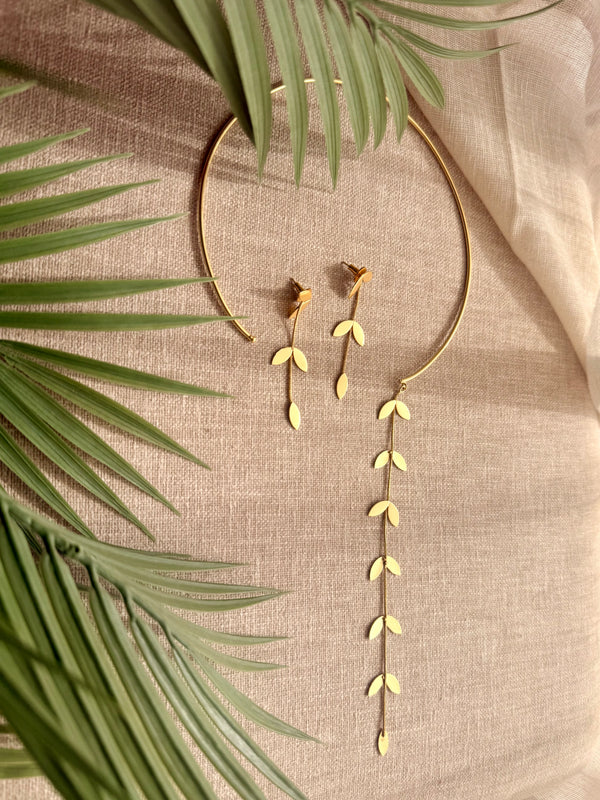 Pallavi set flatlay on grey linen with dried palm fronds — gold-plated wire choker with trailing leaf vine pendant and matching leaf drop earrings, soft natural light