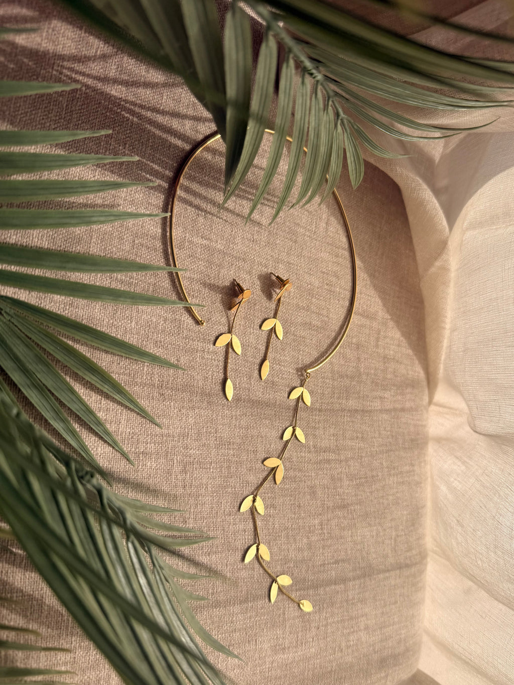 Close-up flatlay of Pallavi earrings resting against the choker, showing cascading leaf-on-chain detail, palm frond in background on linen fabric