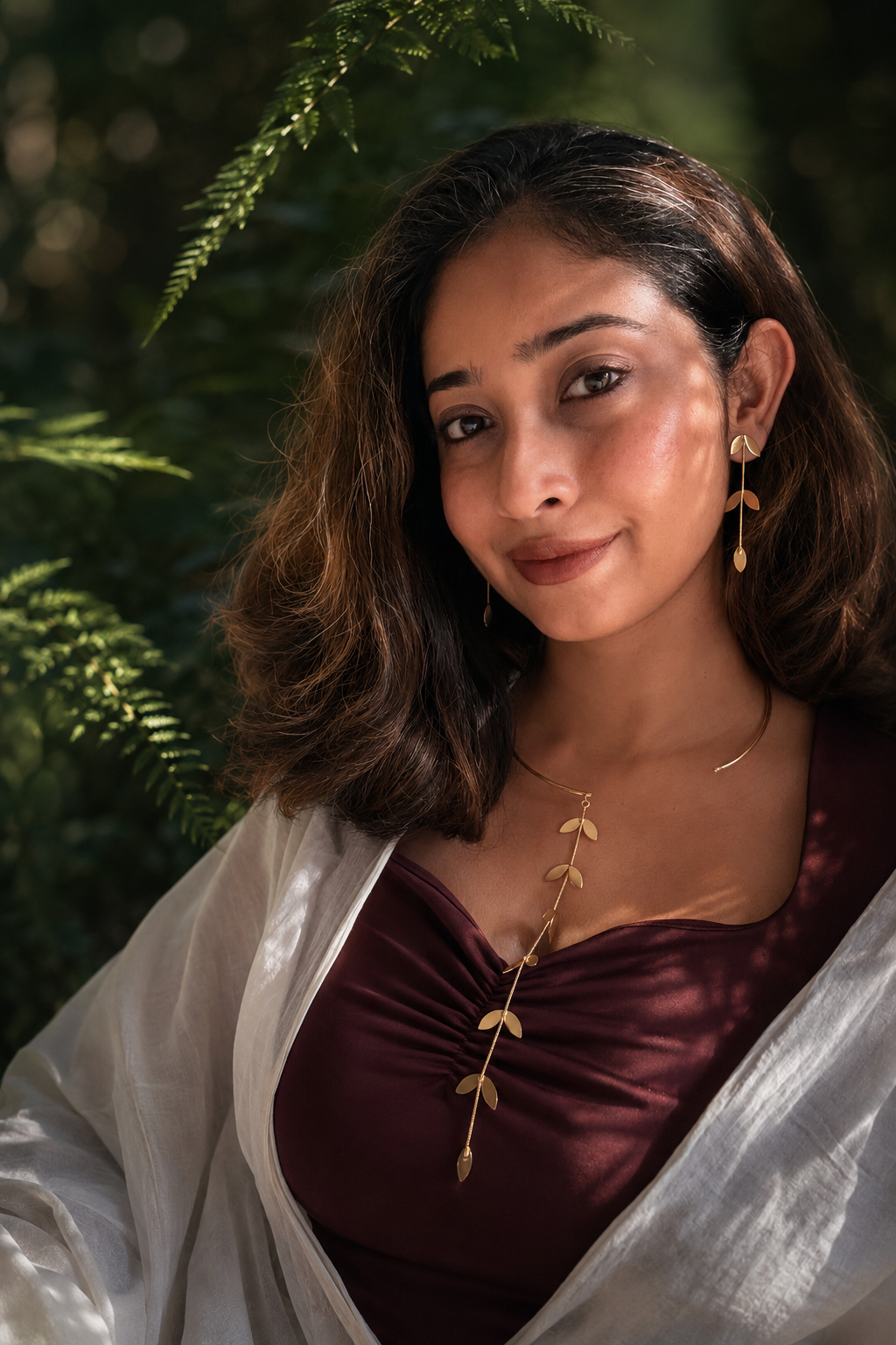 Woman wearing Pallavi set styled with vine centred on choker, dark maroon outfit, lush green botanical background, warm natural light