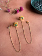 A flat lay photograph of a pair of gold Panna Studs with Chain on a terracotta background next to pink flowers. The earrings feature geometric hexagonal studs set with green stones and a central pearl, each attached to a long gold link chain that ends in a hook detailed with tiny pearls.