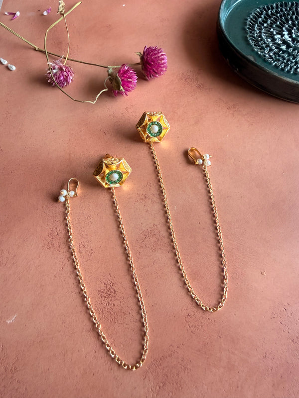 A flat lay photograph of a pair of gold Panna Studs with Chain on a terracotta background next to pink flowers. The earrings feature geometric hexagonal studs set with green stones and a central pearl, each attached to a long gold link chain that ends in a hook detailed with tiny pearls.