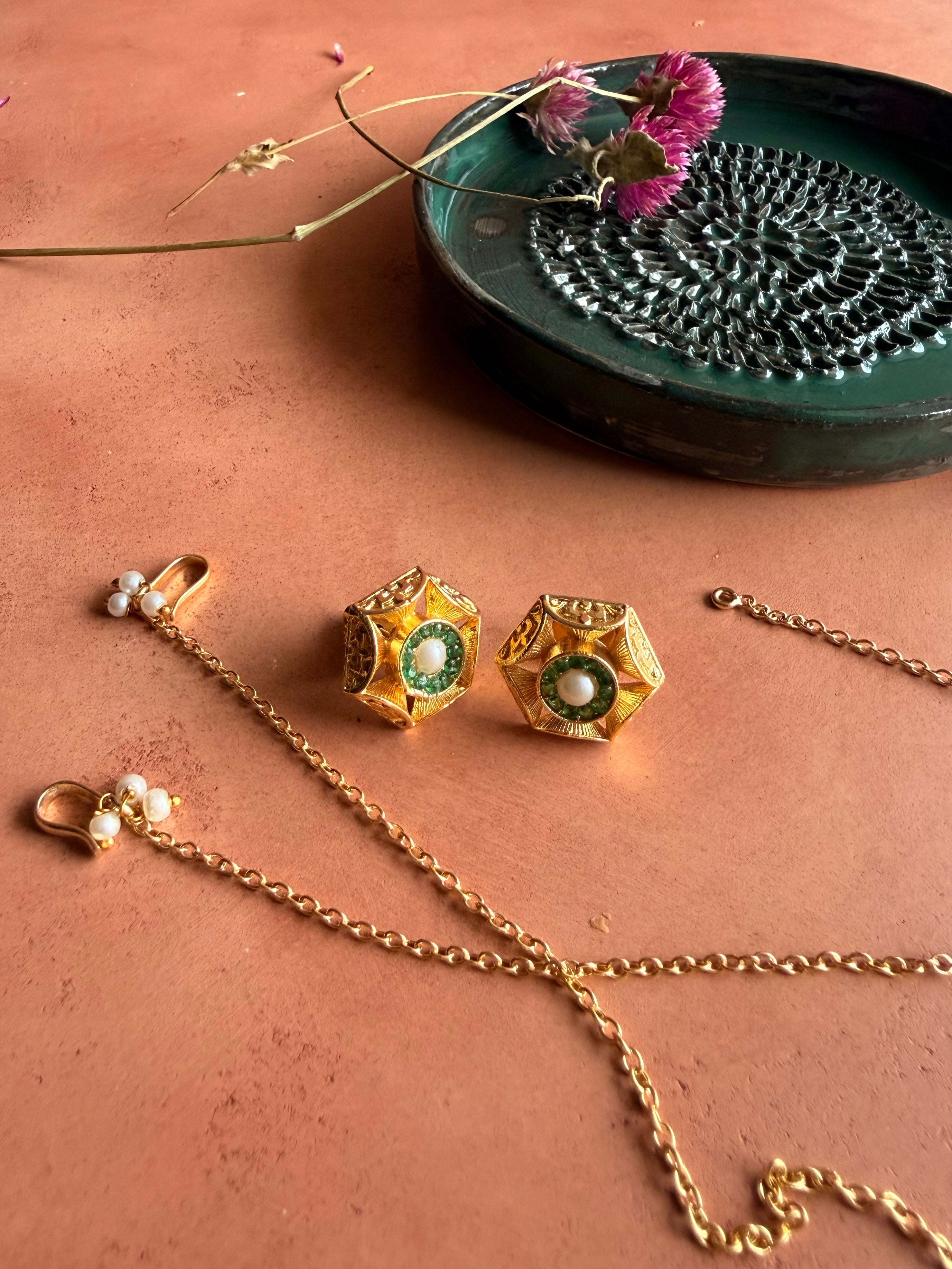 A close-up product shot of a pair of gold hexagonal earrings with green stone halos and central pearls, resting on a terracotta surface. The separate delicate gold ear chains with pearl-trimmed hooks lie gracefully beside them.