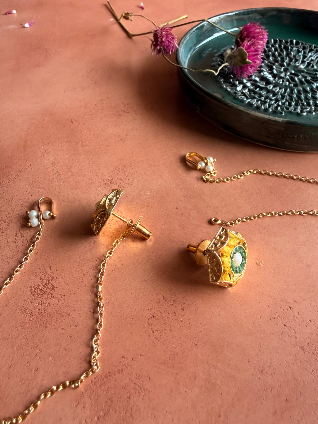A detailed product photograph showing the back construction of the gold Panna Studs on a terracotta surface. One earring is turned to display its traditional screw-back post, showing how the gold chain connects to the piece.