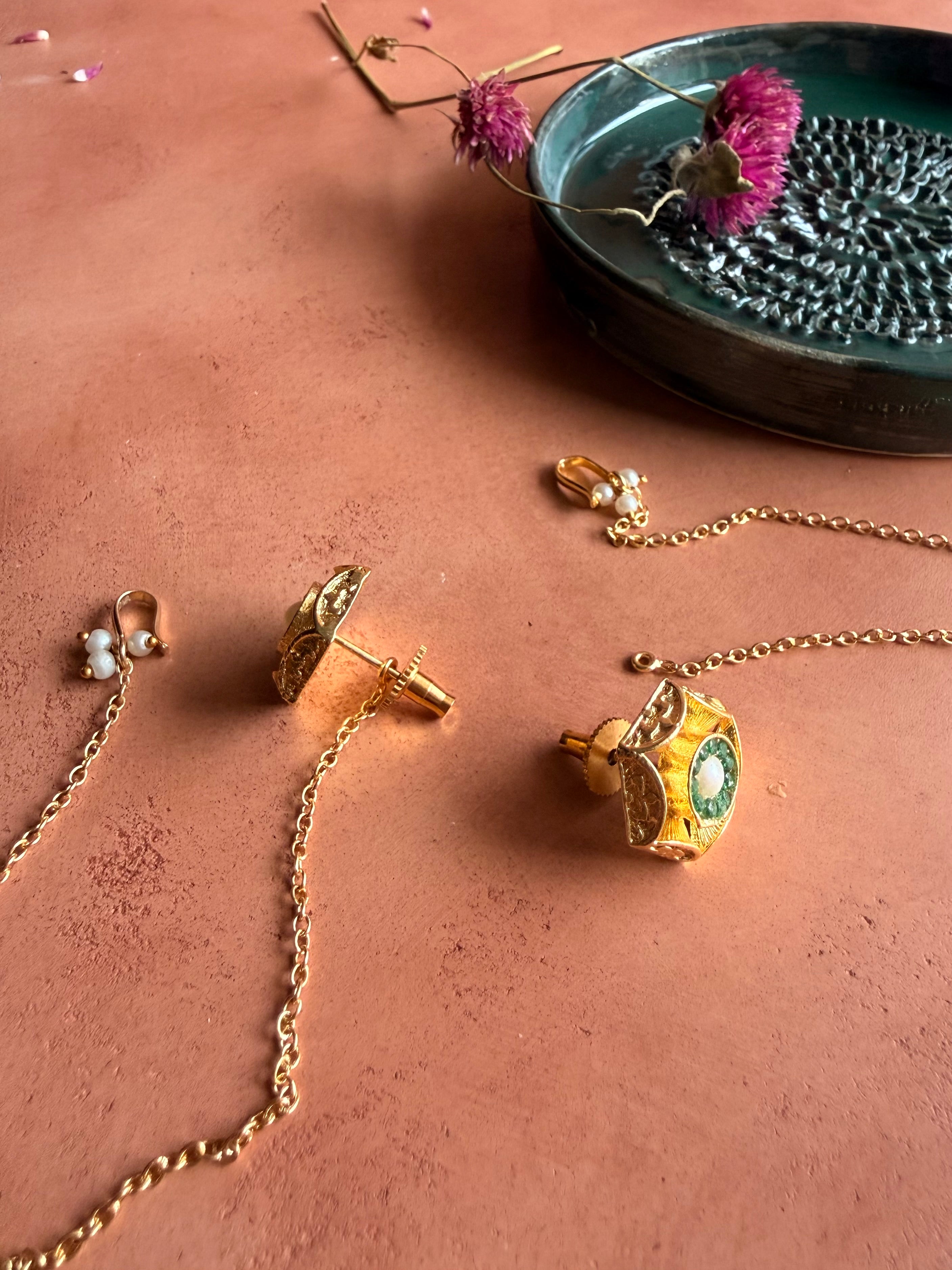 A detailed product photograph showing the back construction of the gold Panna Studs on a terracotta surface. One earring is turned to display its traditional screw-back post, showing how the gold chain connects to the piece.