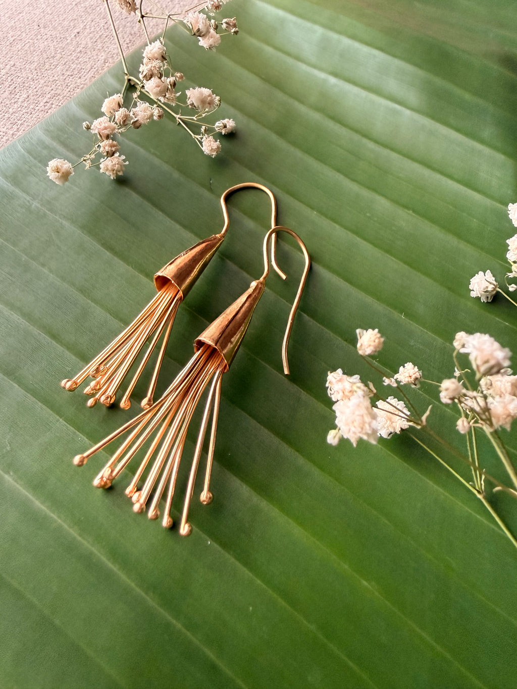 Close-up flat-lay of gold Parag Dangler earrings styled on a fresh green leaf alongside delicate white baby's breath flowers.