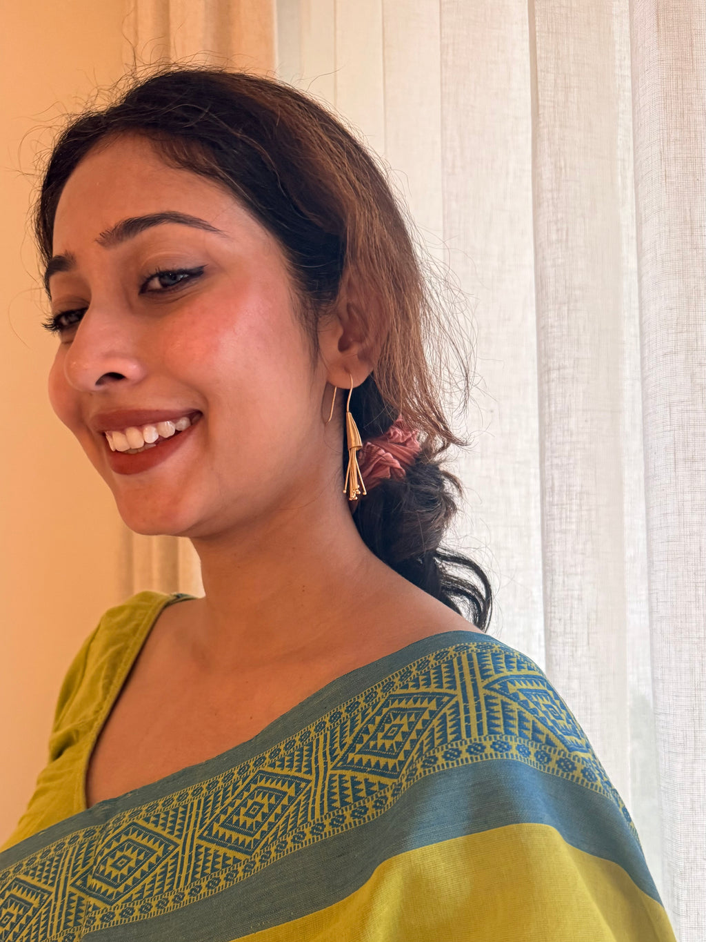 Woman smiling and wearing the gold Parag Dangler earrings, styled with a lime green saree with blue border and a low hair bun.