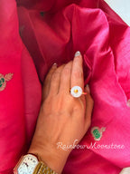 Rainbow Moonstone ring, a round milky white stone with a small gold dot, worn on a hand resting on pink silk