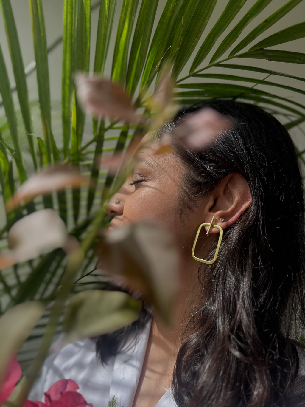 Woman wearing a gold toned rectangular earring with plants in the background