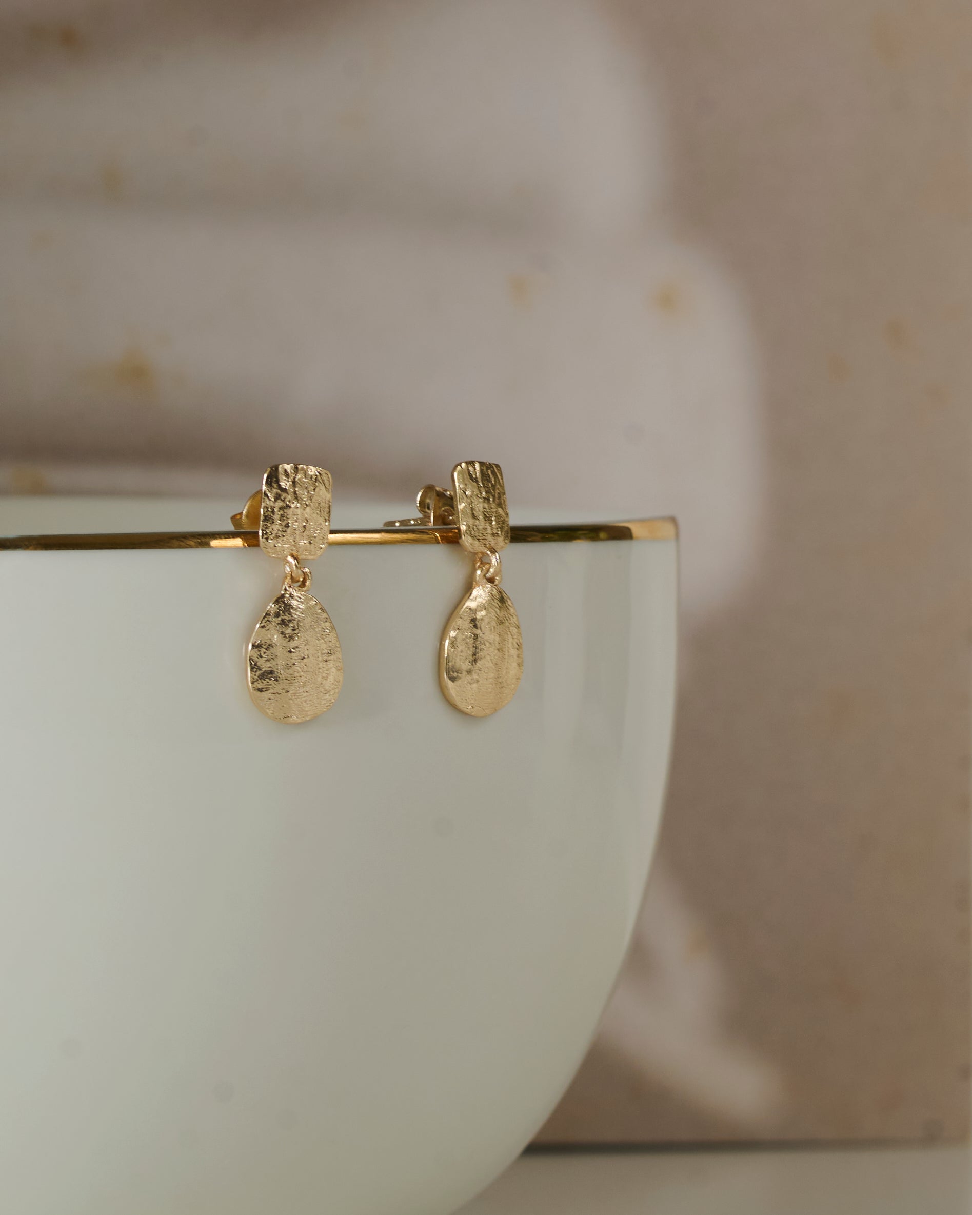 Close up of Small textured gold toned earrings with rain drop shape danglers kept on a white mug.