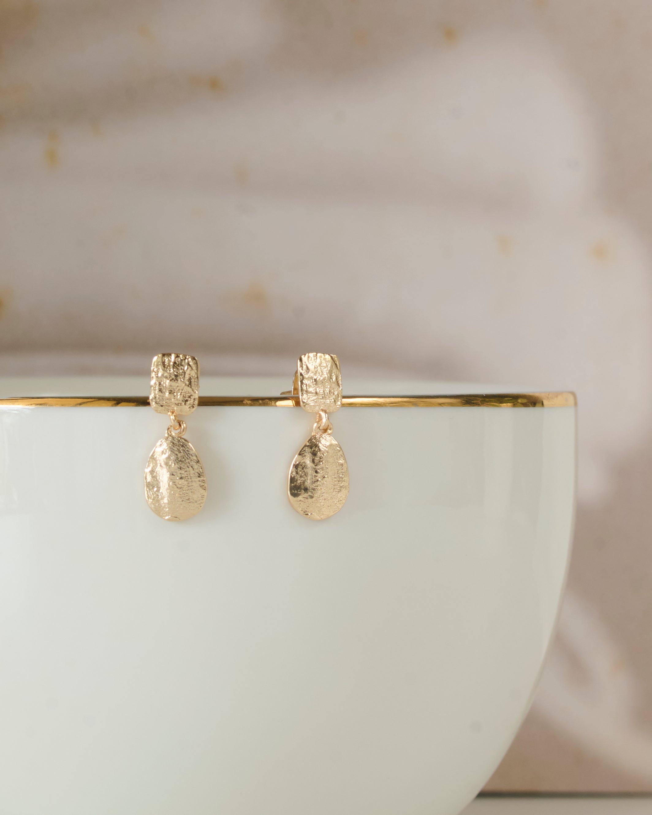 Front view of gold toned earrings with rain drop shape danglers kept on a white mug.