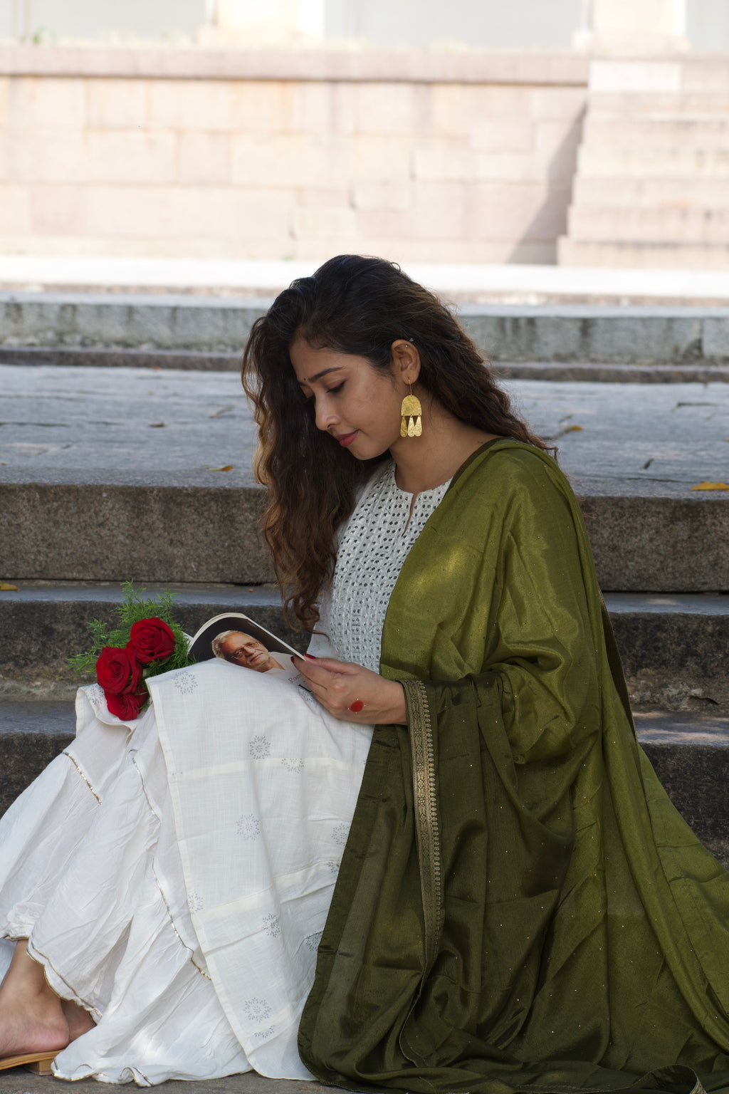 Woman holding a poetry book with red roses against a neutral background, wearing dangling gold earrings that catching the sunlight.
