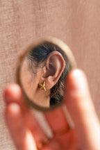 Close-up through a round mirror of a woman's ear wearing a small gold starfish stud earring, soft blurred coral-pink background.