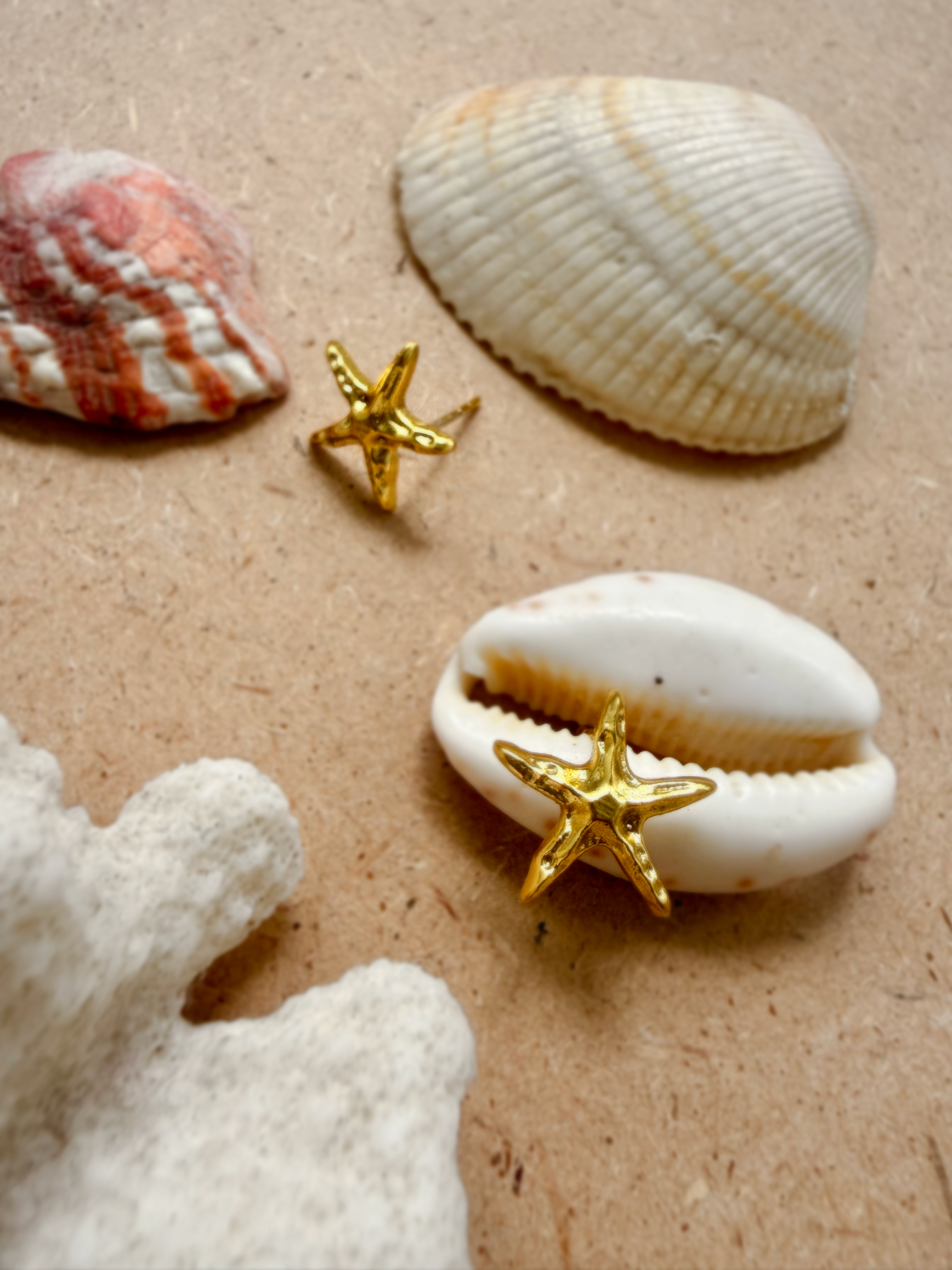 Two gold starfish stud earrings (Sagar Studs) resting on a sandy surface among seashells and white coral.
