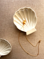 A gold shell-shaped pendant necklace with a delicate chain, displayed inside a white ceramic shell-shaped dish on a textured beige linen background.