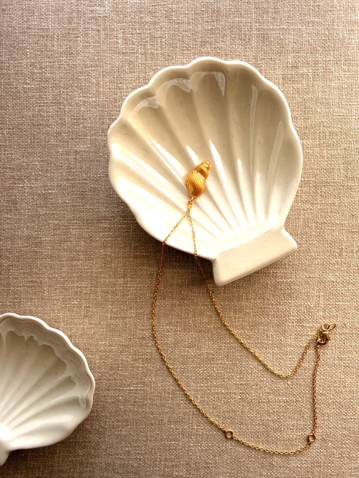 A gold shell-shaped pendant necklace with a delicate chain, displayed inside a white ceramic shell-shaped dish on a textured beige linen background.