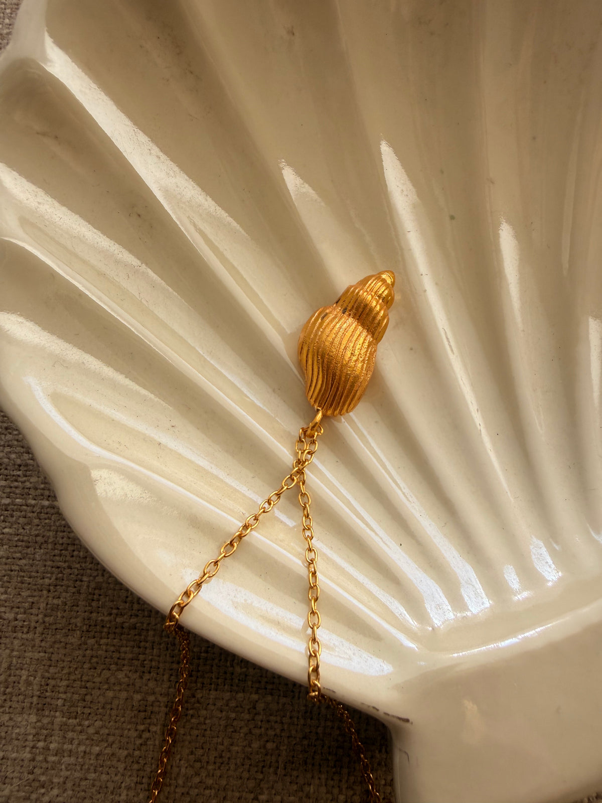 A gold-toned pendant necklace shaped like a ribbed seashell, resting on a glossy white ceramic seashell-shaped dish over a brown textured fabric background.