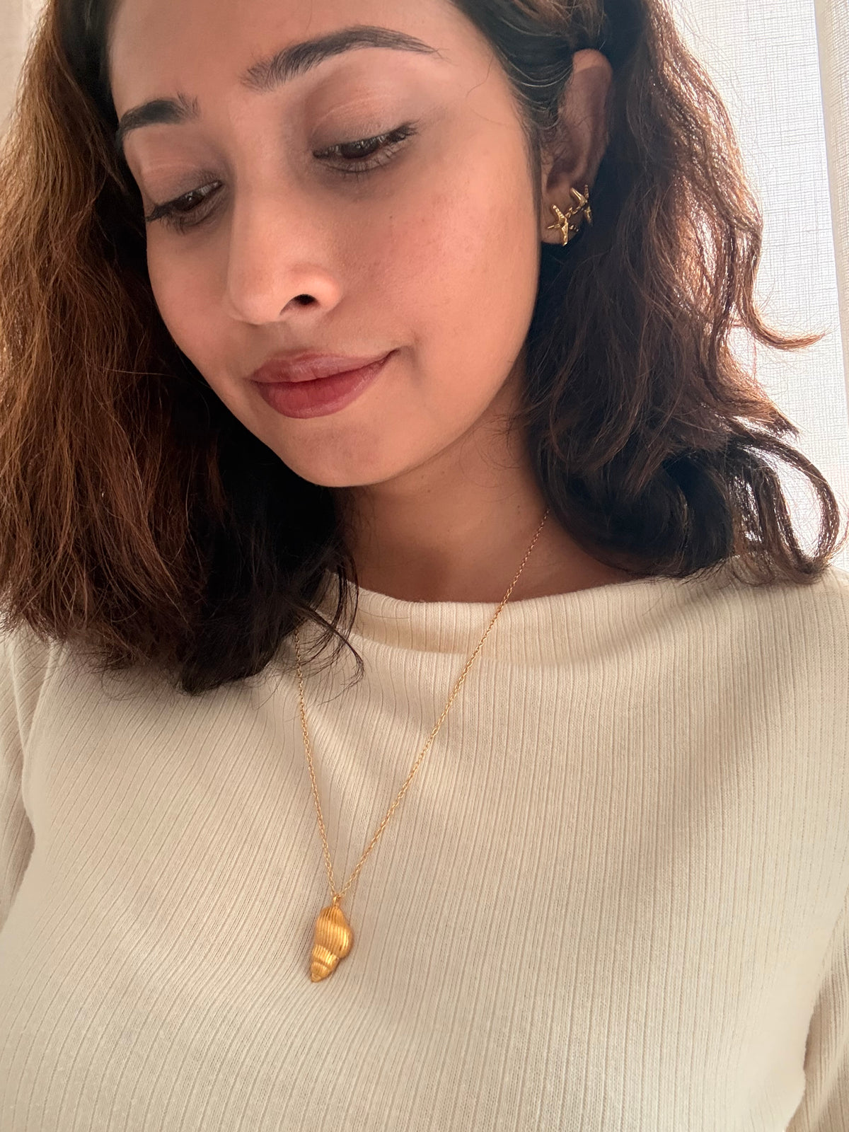 A woman wearing a delicate gold chain necklace with a small seashell-shaped pendant, paired with a cream ribbed top.