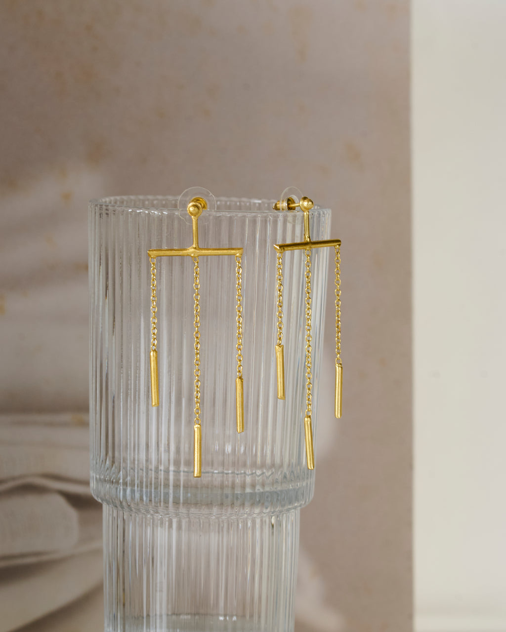 Fluted glass vase with gold toned chain dangling earrings on neutral background.