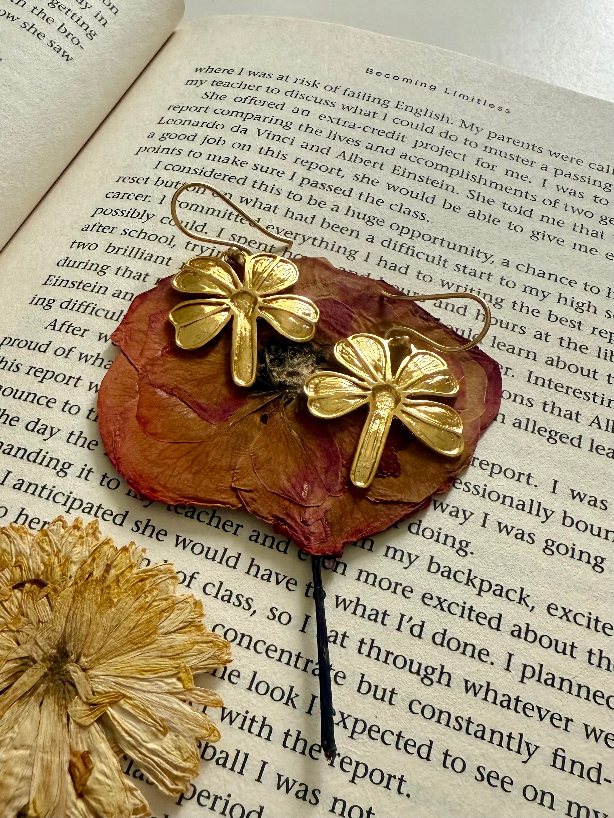 Close up of gold plated four-leaf clover earrings on a red pressed flower, kept on an open book.