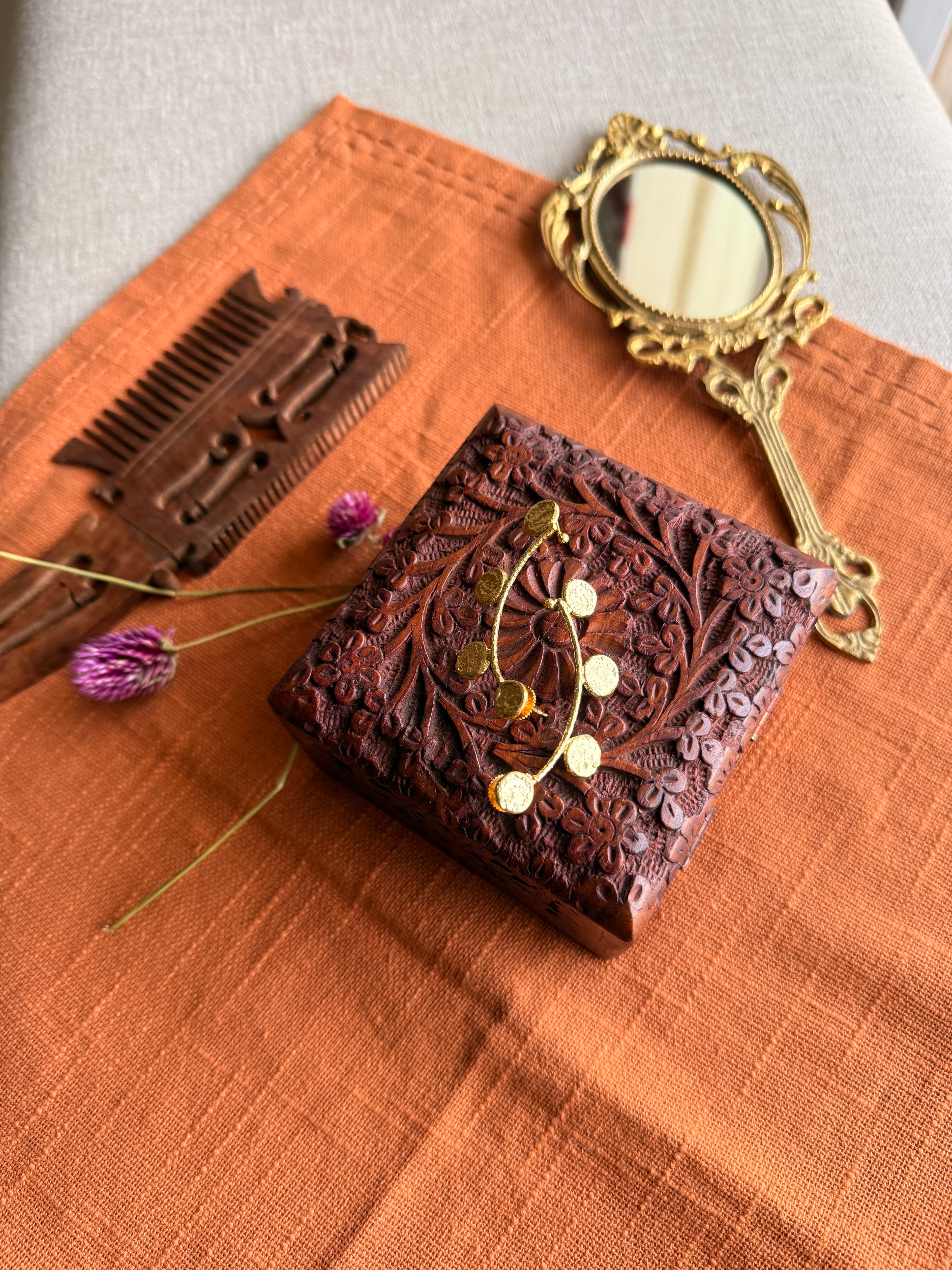 Sitara ear cuffs flatlay on terracotta fabric alongside a carved wooden box, antique hand mirror, and dried purple Globe Amaranth flowers, warm natural light