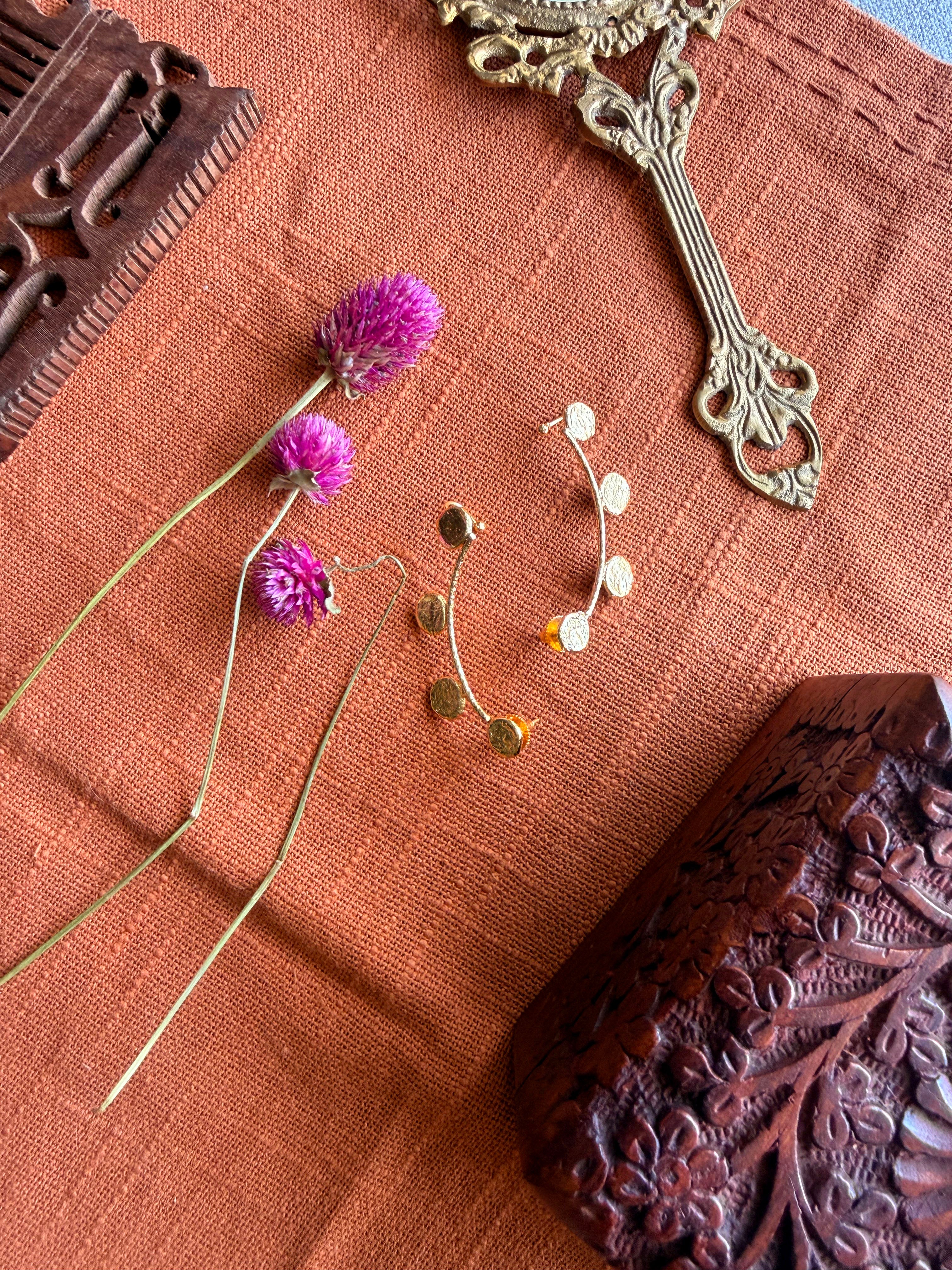 Sitara ear cuffs flatlay on terracotta linen showing both cuffs separated, styled with dried pink and purple flowers, carved wooden box and antique mirror handle in background