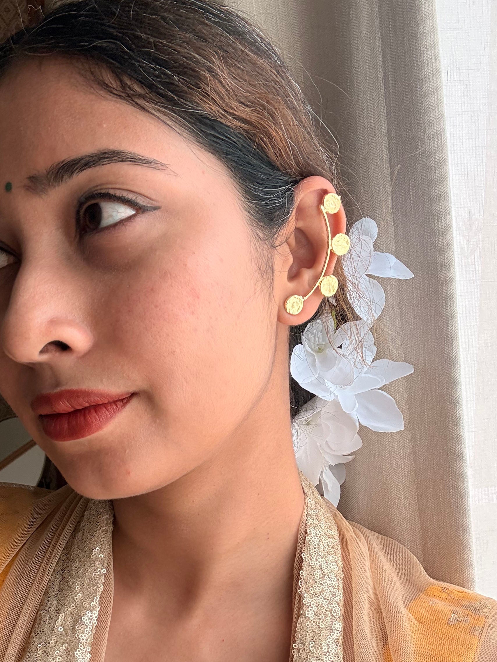 Side profile of woman wearing Sitara ear cuffs, coin disc arc visible curving up the ear, white flowers tucked in hair, golden outfit, natural window light
