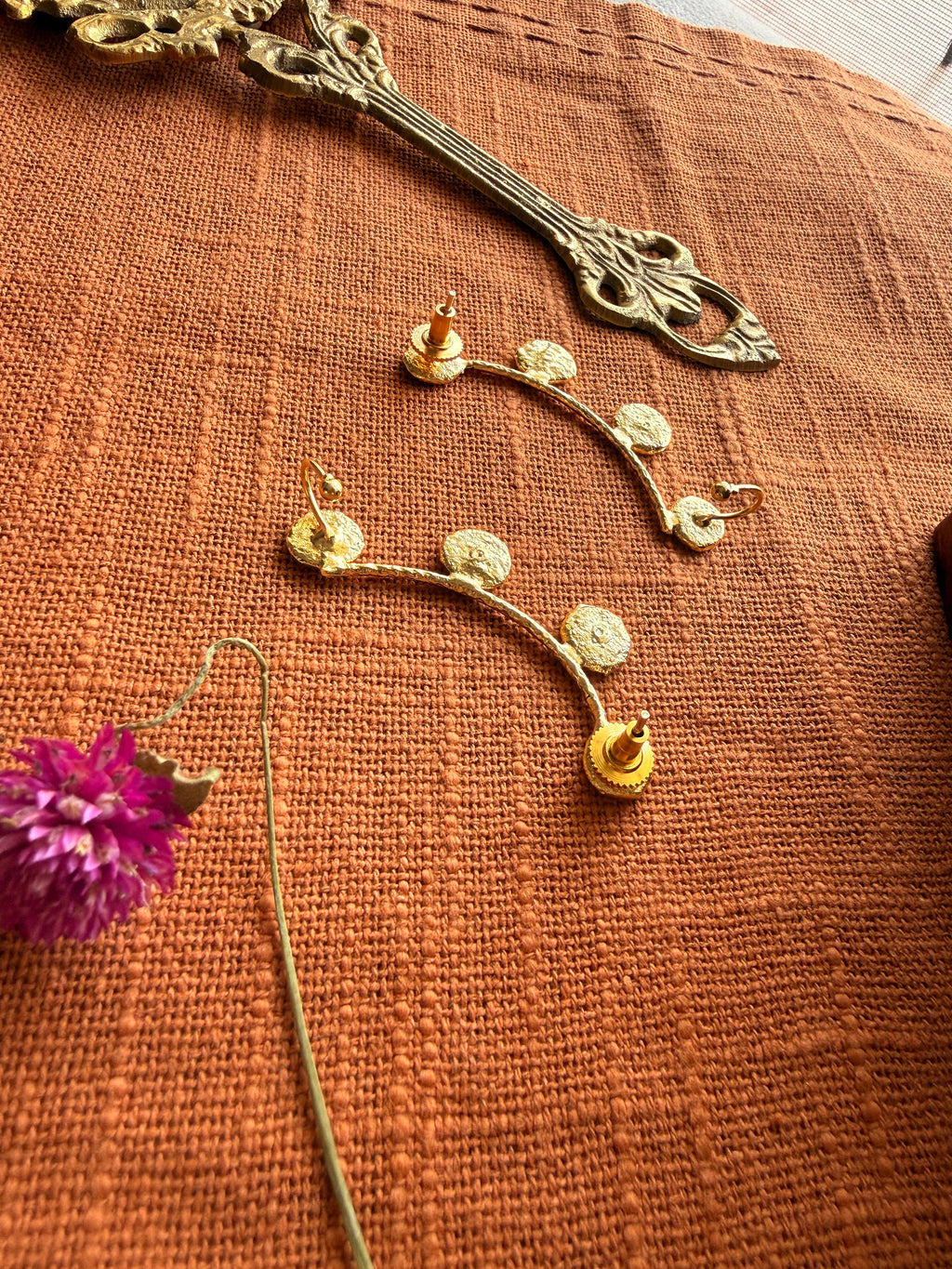 Close-up flatlay of Sitara ear cuffs back side on terracotta fabric, coin disc detail clearly visible, dried purple Globe Amaranth flower beside them