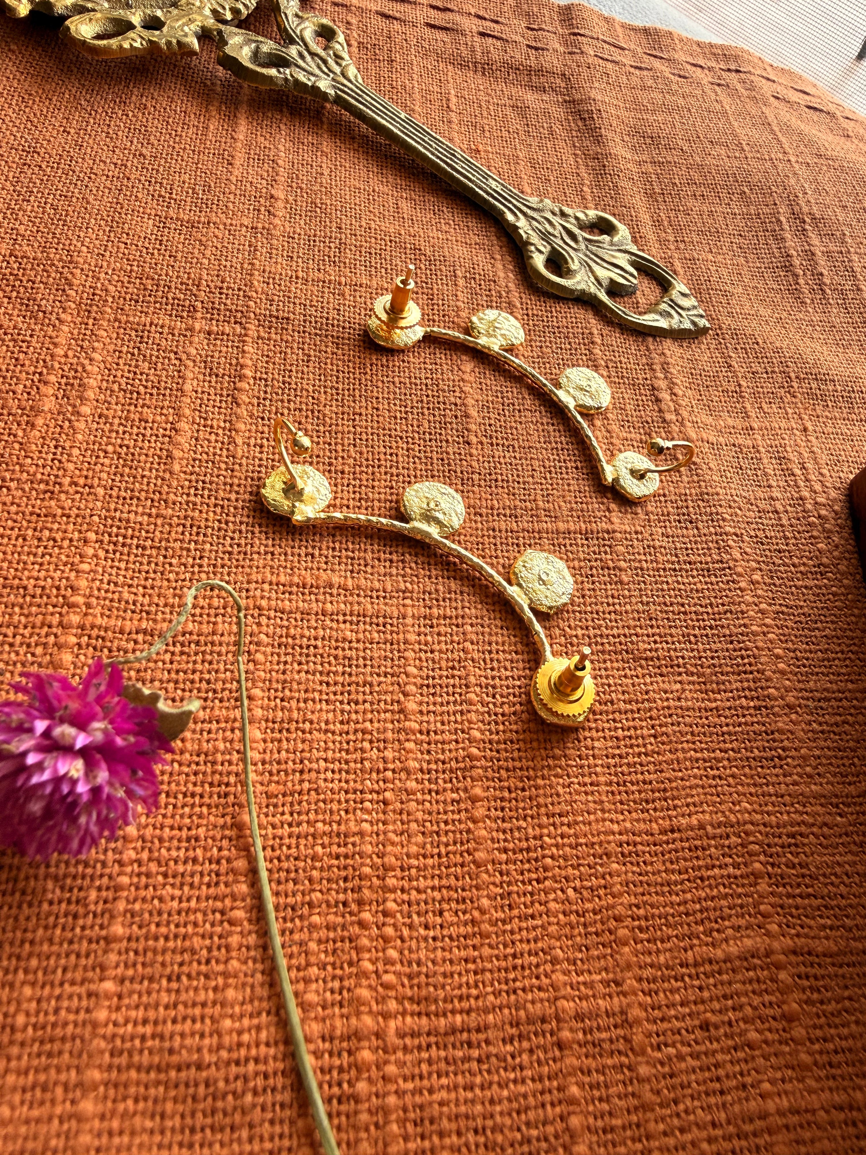 Close-up flatlay of Sitara ear cuffs back side on terracotta fabric, coin disc detail clearly visible, dried purple Globe Amaranth flower beside them
