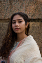 Woman wearing a white kurta with a patterned border against a textured wall, wearing gold plated geometric ear studs.