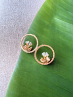 Top-down flat-lay shot of a pair of gold Tagar Stud earrings featuring a pearl-tipped flower bloom inside a textured circular halo, resting on a green leaf.