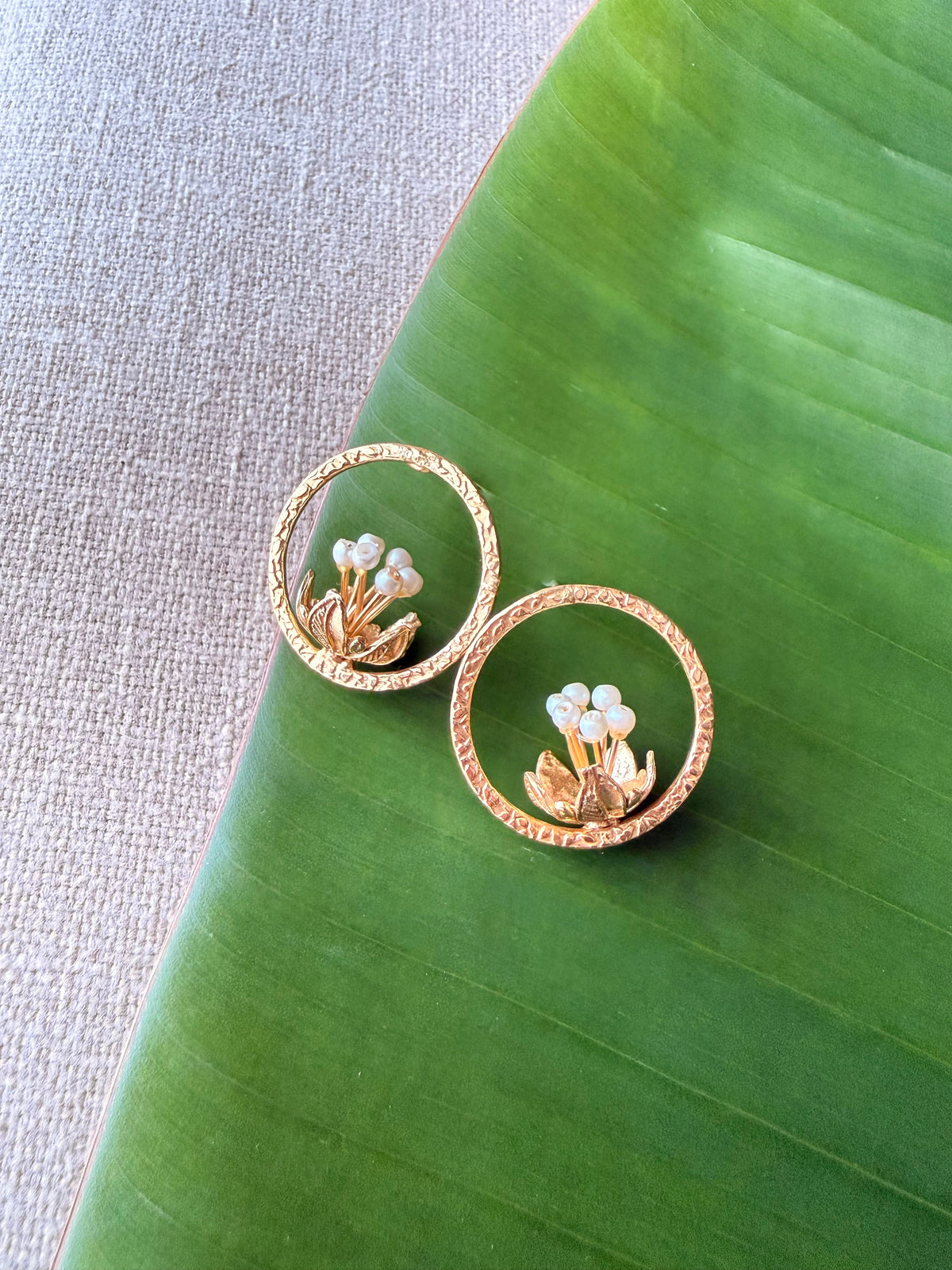 Top-down flat-lay shot of a pair of gold Tagar Stud earrings featuring a pearl-tipped flower bloom inside a textured circular halo, resting on a green leaf.