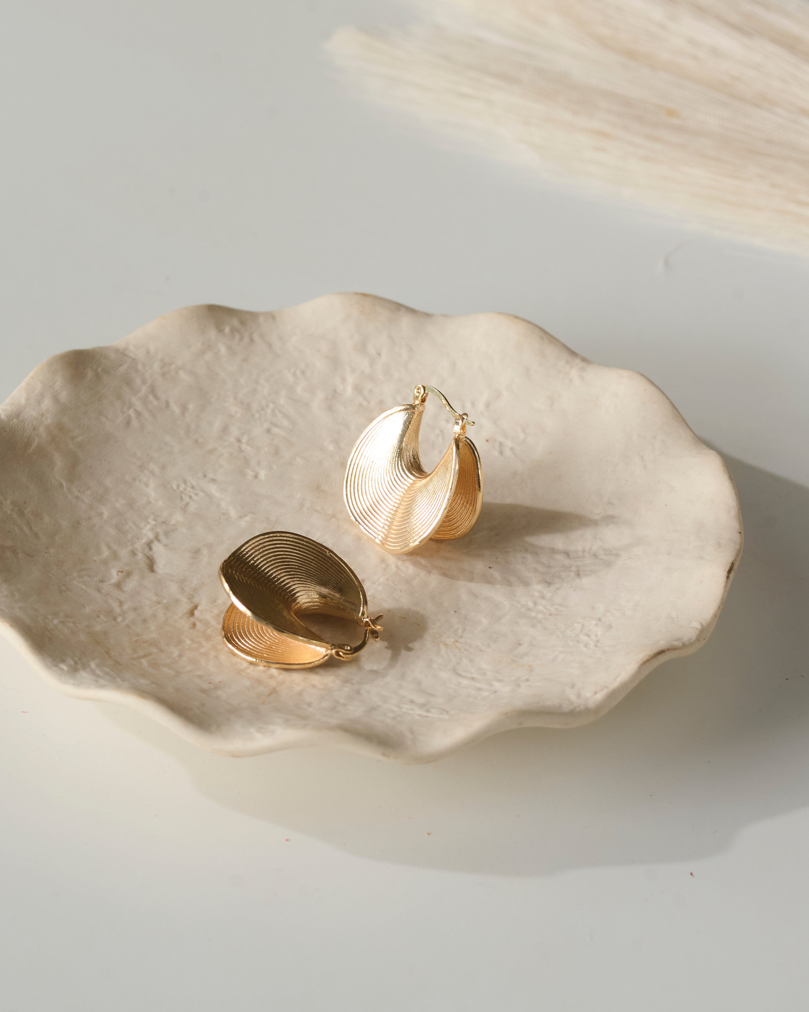 two angle views of Gold hoop earrings with ripple effect design on a textured beige plate with scallops