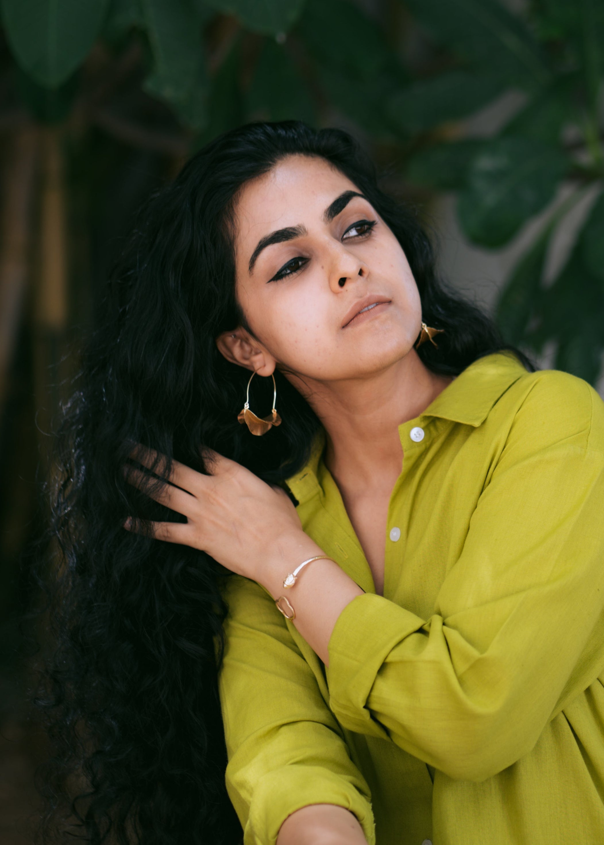 Woman with long dark hair wearing a green shirt against a leafy background, wearing Tarang Leaf Hoops.