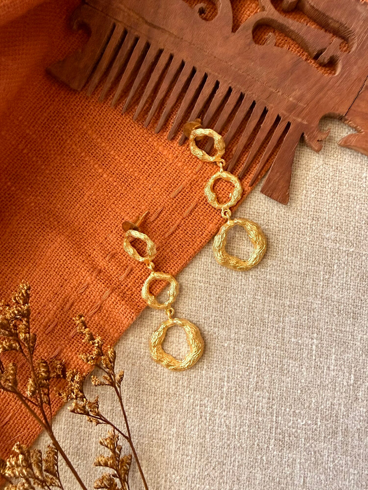 Valaya Danglers laid flat on rust and beige linen, three linked textured gold rings per earring, beside a carved wooden comb and dried flowers