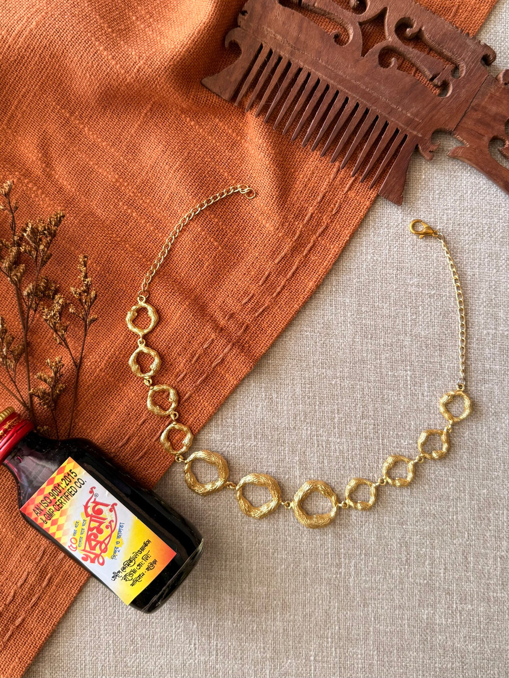 Valaya Necklace laid flat in a curve on beige linen, a row of linked textured gold rings with a clasp and chain, beside a carved wooden comb, dried flowers and a small glass bottle on rust cloth