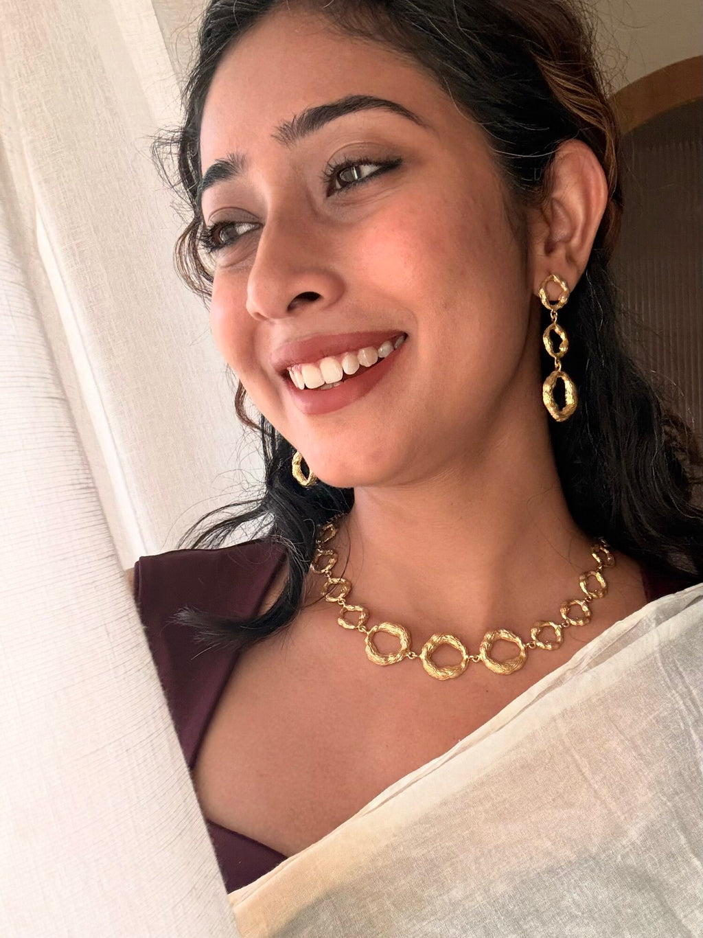 Woman smiling by a sheer curtain wearing the Valaya Necklace at the collarbone with Valaya Danglers, white saree and maroon blouse