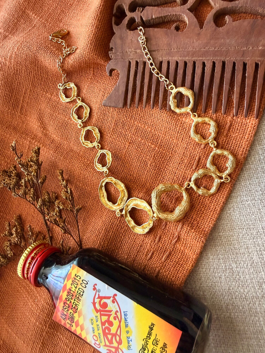 Valaya Necklace flat lay on rust cloth showing the linked textured rings and adjustable chain, beside a carved wooden comb, dried flowers and a glass bottle